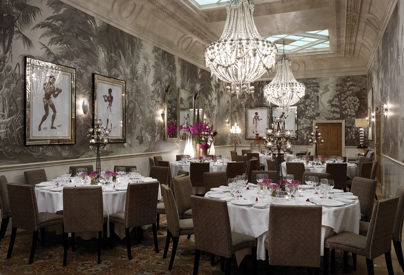 The Shooting Gallery at Haymarket Hotel.  A grand room with high ceilings. A dark grey wallpaper patterned with tropical plants with ornate chandeliers hanging from the roof.  Five round tables are laid up for a formal dinner.