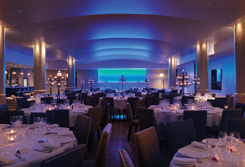 The pool at Haymarket Hotel set up for a dinner party. Several large round tables are laid up for a formal dinner party. In the background is a blue light instillation.