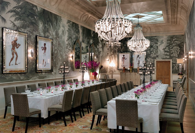 The Shooting Gallery at Haymarket Hotel with dramatic high ceilings and graphic grey wallpaper with forest mural. Two long white tables covered in a white table cloth and set for a formal dinner service fill the centre of the room.