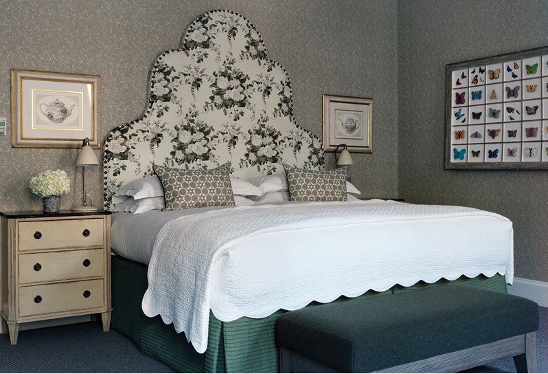A light bedroom with a grey patterned wallpaper, a grey and white floral headboard and forest green end of bed footstool. Two tea cup prints are on either side of the bed and there is an art piece with butterflies.