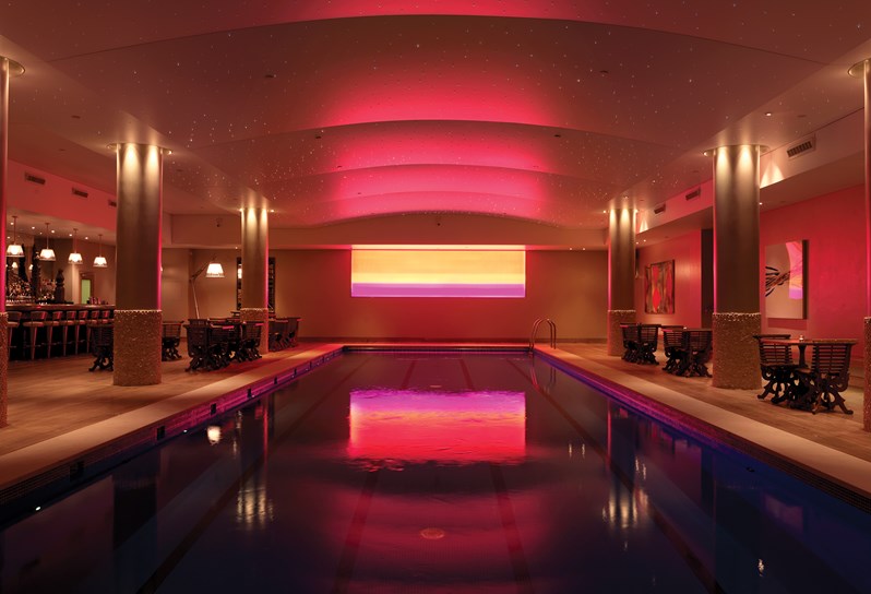 The swimming pool at Haymarket Hotel. An empty indoor swimming pool with an arched ceiling. On the back wall is a pink light instillation.