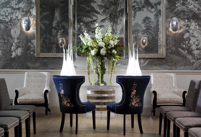 The Shooting Gallery at Haymarket Hotel with dramatic high ceilings and graphic grey wallpaper with forest mural. In the centre of the rooms two blue velvet chairs face a large white flower arrangement.