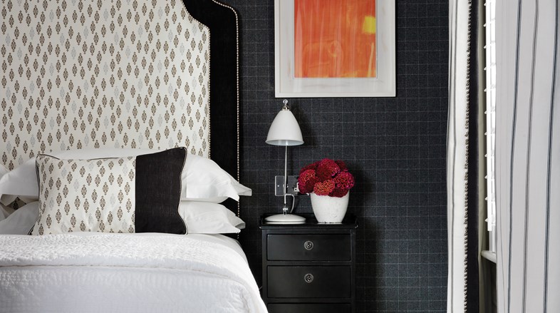 A close up of a magnificent headboard with patterns in white grey and black topping off an inviting king sized bed, placed next to a nightstand table with flowers and original artwork.