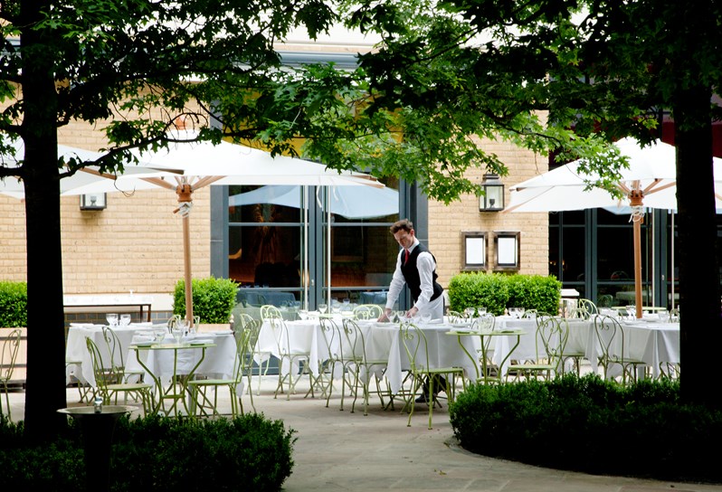 A waiter setting the outside terrace of Ham Yard Restaurant for alfresco dining in the sunshine, with large, leafy trees in the foreground.