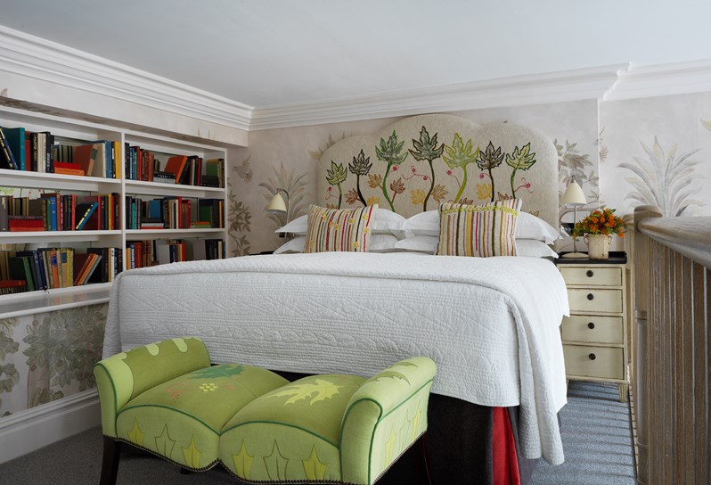 Bedroom area of a mezzanine apartment. The left wall is made up of a bookcase and there is a green ottoman at the end of the bed.