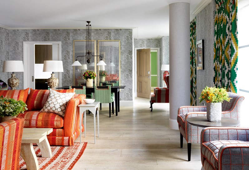 A light spacious living and dining space.  There is a living area to the left with patterned, tonal orange sofas and an oak coffee table. To the right in front of floor to ceiling windows are two grey and orange armchairs and an occasional table with flowers.  
Behind the sofas is a black dining table with four green and white dining chairs.  To the right of this is a orange and grey wingback chair which sits at the top of the short hallway leading to the front door.