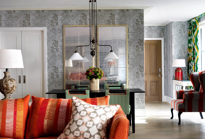 A light and spacious room with light grey patterned walls. There is a patterned orange sofa with a grey and white cushion. There is a large table lamp to the left just behind the sofa.   There is a black dining table with four green and white chairs and a big rectangle antique mirror on the wall.   There are double doors leading to the kitchen to the left of the dining table and an orange and grey wingback chair to the right of the dining table.