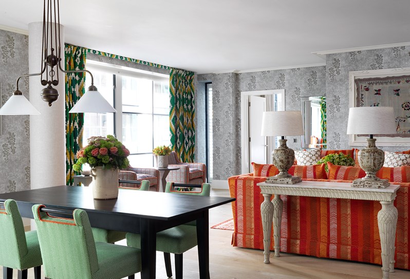 A black dining table with four green and white chairs. Pendant lights hang above the table. Behind the table is a long occasional table with two lamps which sits against the back of an orange patterned sofa. To the left of the sofa are two arm chairs and a table which sit in front of large floor to ceiling windows. The windows have floor length green and yellow patterned curtains. The walls are light grey and patterned.