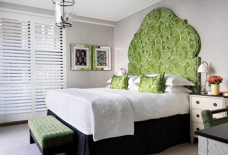 A light and airy bedroom with king-sized bed. The bed has white linen and a large green and white patterned feature headboard. There is a green fabric footstool at the end of the bed, a chest of drawers as a side table, a desk with chair and floor to ceiling windows with white shutters.