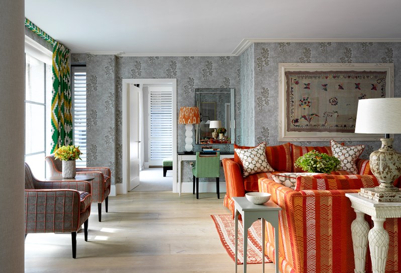 A view of the living area towards the bedroom. A light and airy room with floor to ceiling windows and light grey patterned walls. To the right there are two orange patterned sofas facing each other with side tables and a coffee table.  There is a writing desk and lamp and two grey and orange arm chairs with a table which sit to the left in front of large floor to ceiling windows. The windows have floor length green and yellow patterned curtains.