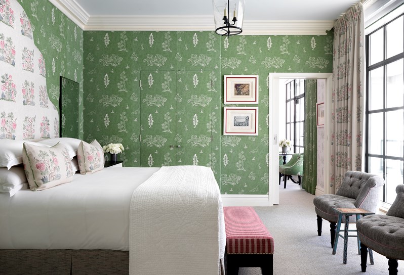 Green bedroom in tones of cream and greem, boasting a king size bed and Kit Kemp's statement mannequin, with a pop of color from a fresh white flower arrangement on a side table.