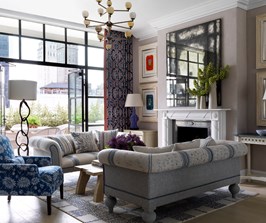 A spacious living room in neutral and blue colors with open French doors leading out to a big terrace. Featuring two inviting sofas, a comfortable arm chair and a wooden coffee table with fresh flowers in front of a fire place.