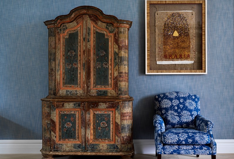 A beautiful antique cabinet next to a bright blue arm chair with white flowers, with original artwork hung above on the denim colored wall.