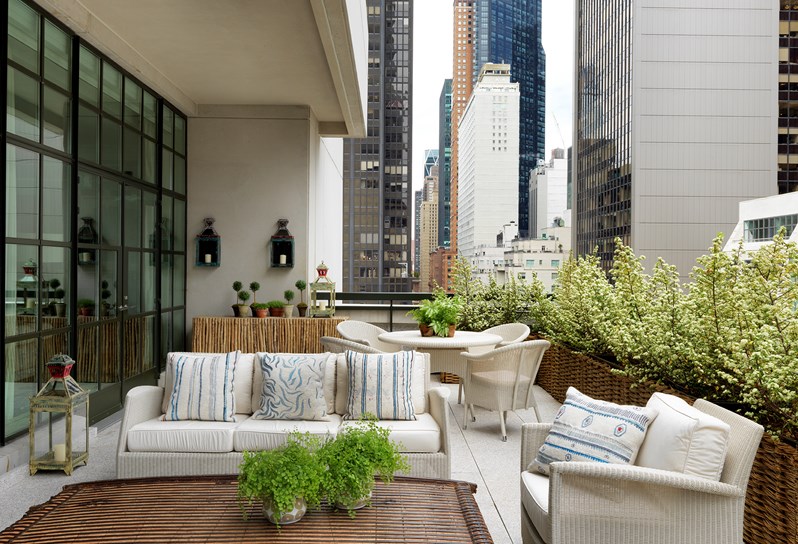 A spacious terrace with beautiful cream patio furniture and sweeping views of Midtown Manhattan, featuring a dining area with a table and four chairs, as well as a lounge area with a sofa and an arm chair, all surrounded by leafy plants.