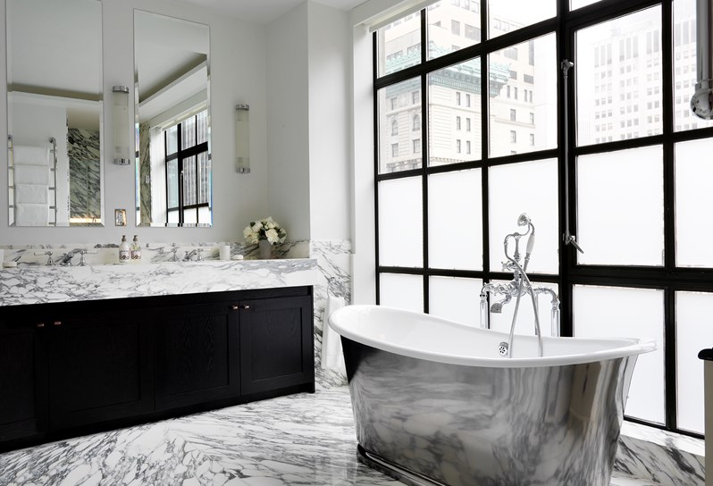 A spacious and airy white marble bathroom with a free standing chrome bathtub in front of frosted floor to ceiling windows, a wash basin with two sinks and over sized mirrors.