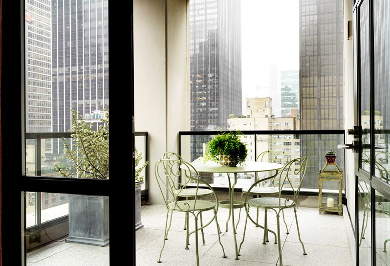 A spacious terrace with views of Manhattan, containing four chairs and a table in summery light green metal, a large floor lantern and leafy plants.