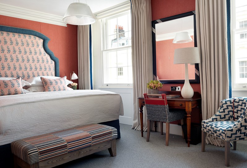 A bedroom in shades of blue, white and salmon with blue trimmed headboard. There is a blue and white chair and cream drapes.
