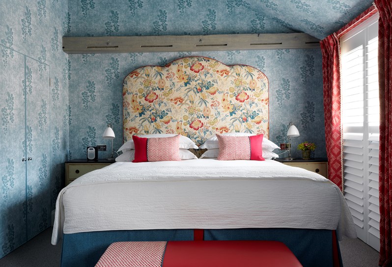 A view of the bed straight on. The room has a blue patterned wallpaper and a floral headboard in yellow, red and blue. To the right is a window with red curtains and there are also pops of red with the decorative pillows and the end of bed footstool.