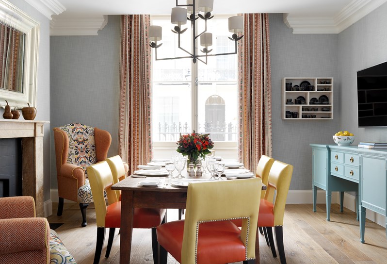 A light and bright dining room with big window in the Townhouse. There is a dining table in the center of the room with yellow and orange chairs. A fireplace is to the left and is framed by two wingback chairs. To the right of the dining table is a blue table with television on the wall.
