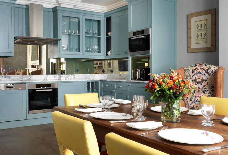 A shot of the kitchen in the Townhouse. In the forefront is the dining table with flowers in the center and yellow and orange chairs. In the background is the kitchen with blue cabinets and beautiful marble countertops.