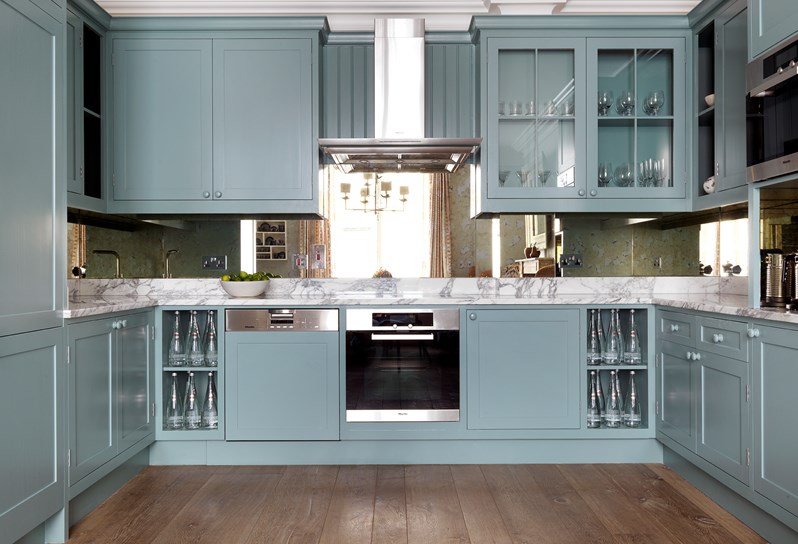 A closer view of the kitchen in the townhouse pictured straight on with the oven and dishwasher in the center. Marble countertops and blue cabinets, some with glassware visible.