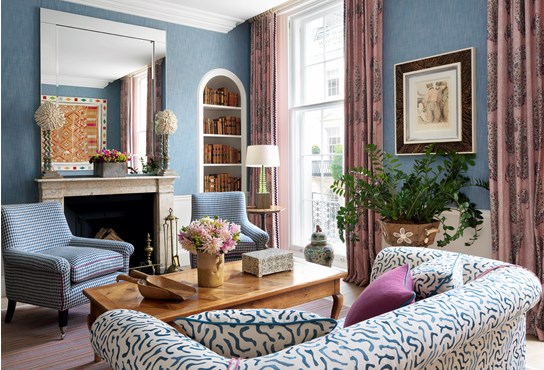 A living room with a fireplace. There are blue walls, a sitting area with blue patterned chairs in front of the fireplace, a coffee table and a blue and white couch in the forefront. There are large windows to the right with maroon drapes and in between the windows is greenery and art work.