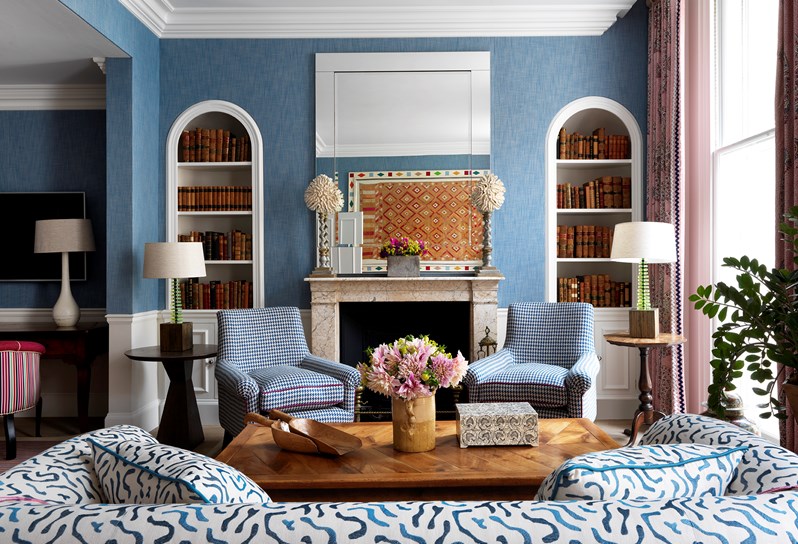 A straight on view of the fireplace in the living room. There are blue walls with a blue and white couch in the forefront. Above the fireplace is a mirror and there are two built in bookcases on either side. In front of the fireplace are two chairs in a blue patterned fabric.