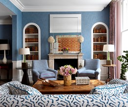 A straight on view of the fireplace in the living room. There are blue walls with a blue and white couch in the forefront. Above the fireplace is a mirror and there are two built in bookcases on either side. In front of the fireplace are two chairs in a blue patterned fabric.