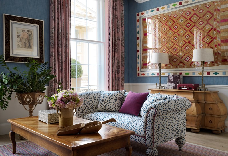 A close up view of a blue and white patterned sofa in the living room. There is a greenery and art work next to the window and in the background a large framed tapestry hangs on the wall above a dresser table.
