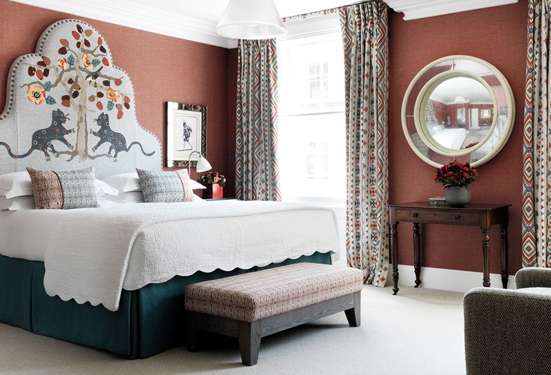 Bedroom with  a whimsically patterned headboard in shades of grey, brown, red, blue and orange. A deep red wallpaper and two side tables with reading lamps. To the right wall is a circular mirror and table in between the two large windows.