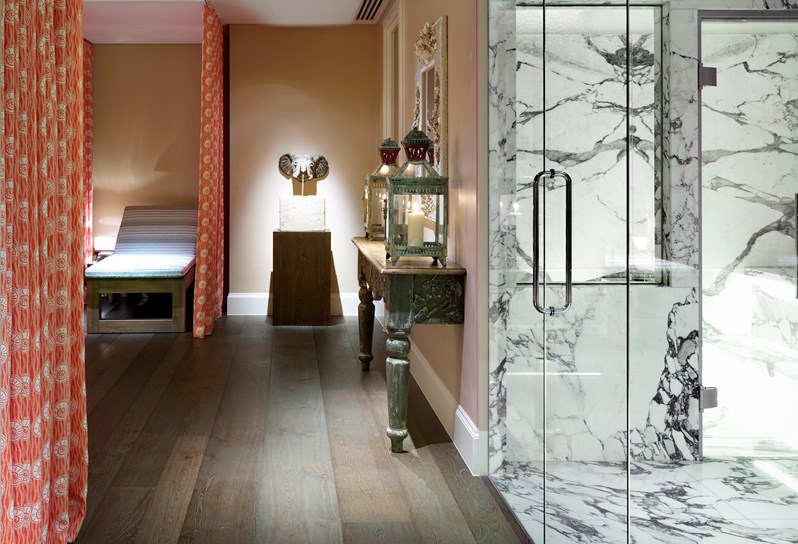 Look through onto the spa relaxation area. There are orange and white curtains hanging from the ceiling and a white relaxation bed. On the right you can see the marble steam room. At the very end of the room there is a small table with an elephant shaped fountain.
