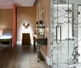 Look through onto the spa relaxation area. There are orange and white curtains hanging from the ceiling and a white relaxation bed. On the right you can see the marble steam room. At the very end of the room there is a small table with an elephant shaped fountain.