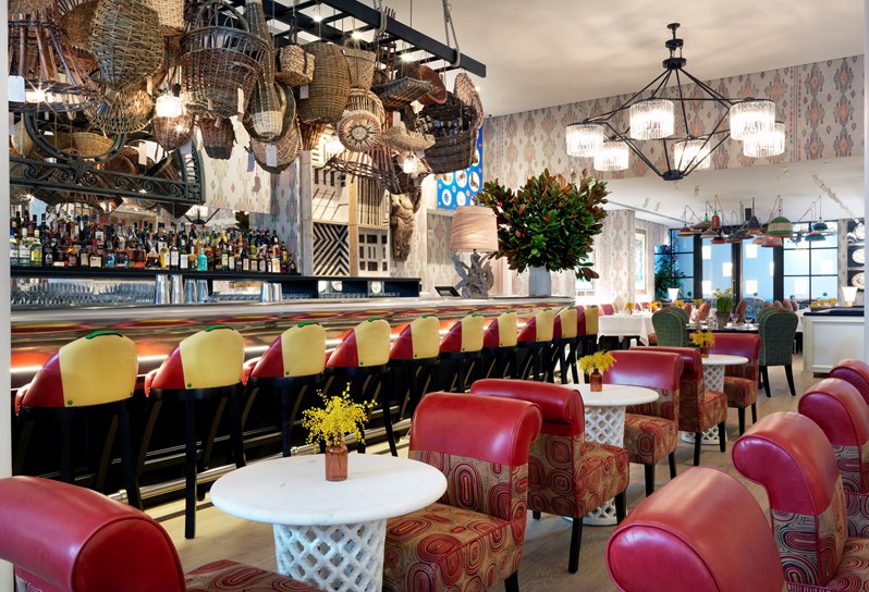 A side view of The Whitby Bar with its art installation of handcrafted British baskets, showing comfortable bar seating at both high chairs by the bar as well as lower lounge furniture in bold colors, all surrounded by stunning flower arrangements and original artwork.