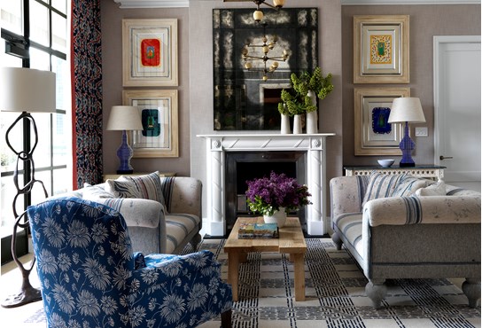 A bright living room in neutral and blue colors with floor to ceiling windows, featuring two inviting sofas, a comfortable arm chair and a wooden coffee table with fresh flowers in front of a fire place, surrounded by original artwork.