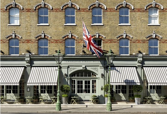 An exterior shot of the Charlotte Street Hotel face on from the other side of the street, showing outdoor terrace, olive trees at each side of the entrance doors and Union Jack blowing in the wind.