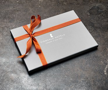 A picture of the actual voucher box that will be received in the post. A grey box with Firmdale Hotels and logo applied, tied with orange ribbon.