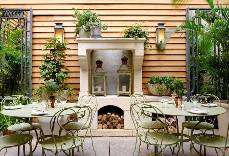 A leafy and airy patio area of the restaurant, containing summery light green metal tables and chairs, as well as lanterns and wooden decorations.