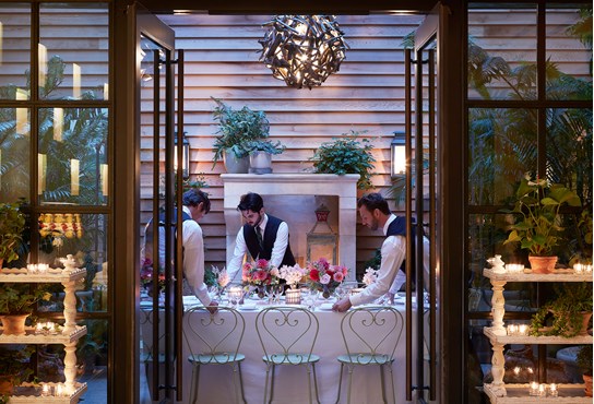 Three members of staff setting up for an evening dinner in the patio area of the restaurant, showcasing a softly lit area with a beautifully set table and several flower arrangements and plants.