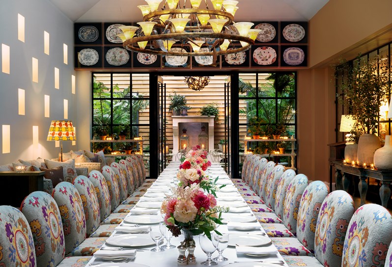 The Orangery set up for private dining for 25 people on one long table with beautiful flower arrangements, with comfortable, padded chairs in patterned fabric around, looking toward the leafy patio area.