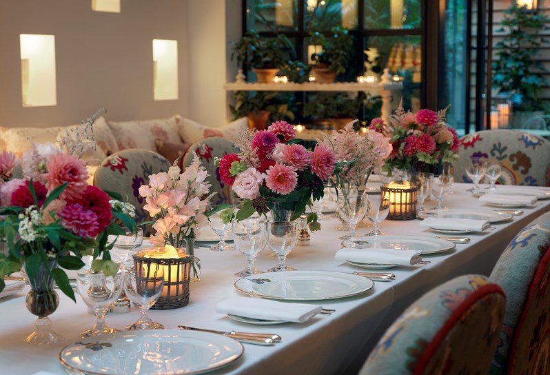 A candlelit table set for dining with Kit Kemp's own Mythical Creatures range for Wedgwood, stunning flower arrangements and comfortable chairs in flowery fabric.
