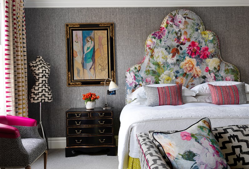 A bedroom with multi coloured headboard, yellow valance and pink and grey cushions on the bed. At the bottom a black and white sofa sits with a cushion matching the bed's headboard. A mannequin is in the far right corner next to a window and bedside chest.