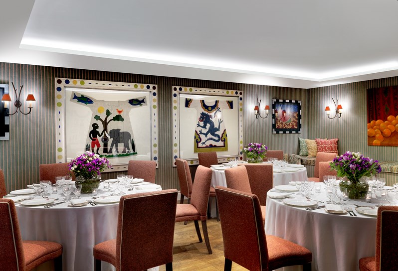The beautiful Vanessa Bell Room setup for a dinner on round tables with orange chairs. There is beautiful bright artwork on the walls and flowers decorating each table.