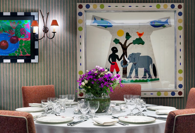 A closeup of a table in the Vanessa Bell Room setup for a dinner orange chairs. There is beautiful bright artwork on the walls and flowers decorating the table.