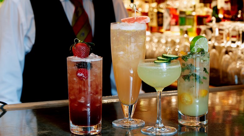 A selection of cocktails on a pewter bar. In background is a bartender in a white shirt
