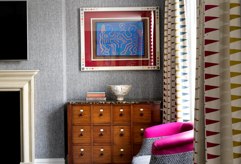 A chest sits in the corner of a room with art above and a grey and cerise tub chair in front. To the right are patterned curtains and to the right is a fireplace and television hanging above.