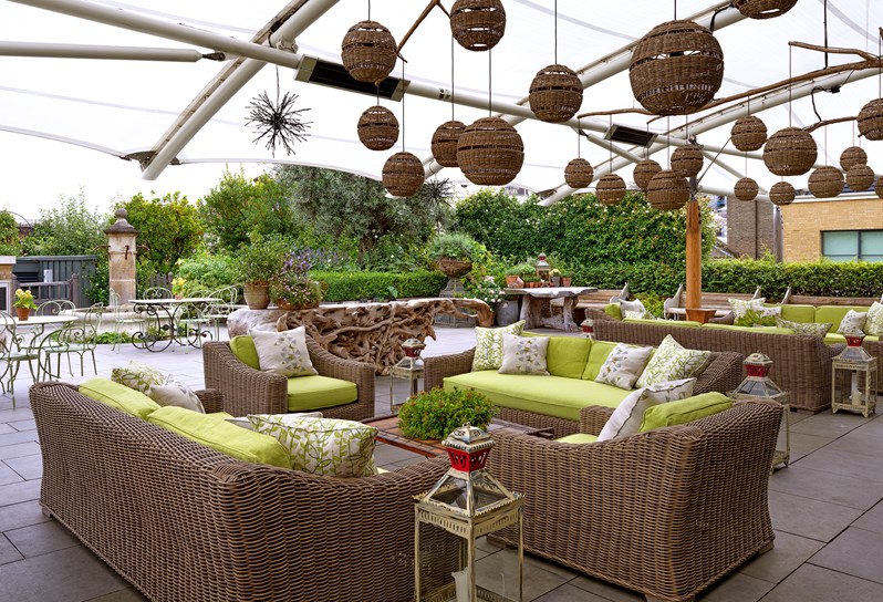 A semi covered, leafy roof terrace; two seating arrangements with rattan sofas and armchairs with  fluffy lime green cushions and coffee tables under a canopy with quirky lanterns and further back an uncovered fountain and leafy garden.