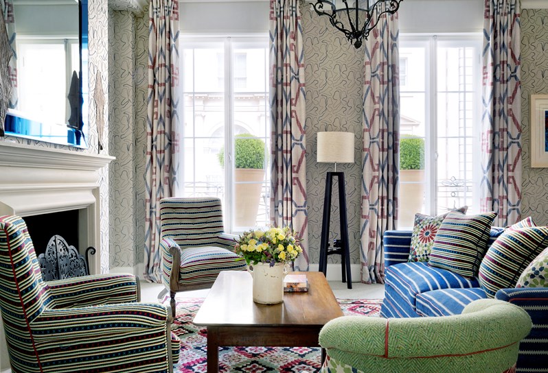 Three small green arm chairs and a large bright blue striped sofa are set around a low wooden coffee table with a vase of yellow flowers. In the background are two floor to ceiling windows, flooding the room with light. To the left of the image is a white wooden fireplace with a blue mirror hanging above.
