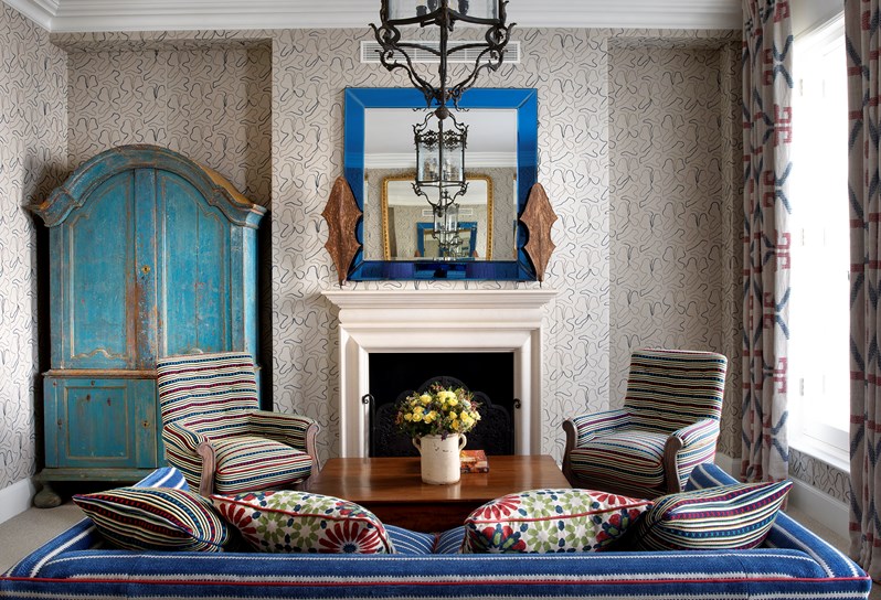 In the foreground is a bright blue sofa. Against the rear wall is a large antique style turquoise wardrobe.  Two small patterned arm chairs are placed around a low dark wood coffee table. In the background is a white fireplace with a bright blue mirror above.