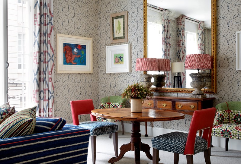 A round mahogany dining room table with blue and red leather chairs.  In the background a dark wooden console table with two stone lamps with red lampshades. Above is a gold gilded mirror and a large turquoise and magenta tapestry hangs on the neighboring wall. The corner of a dark blue sofa can be seen in the foreground.