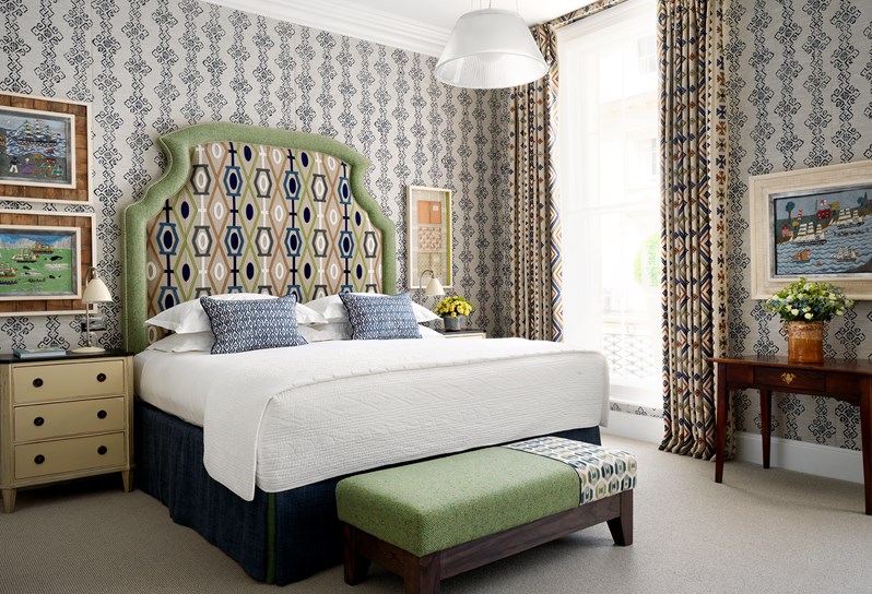A king size bed with a beige graphic headboard sits in front of floor to ceiling Georgian windows. At the foot of the bed is a small pale green footstool.  On the walls are three artworks of sea scenes.